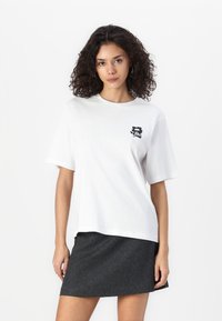 T-shirt en coton blanc avec un col rond, des manches courtes et un graphique noir sur la poitrine gauche. Porté avec une jupe gris foncé.