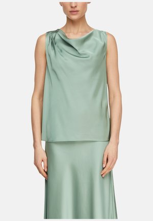 Kobieta ubrana w bezrękawnikową bluzkę z zielonego satynu z marszczonym dekoltem i pasującą do niej spódnicę z wysokim stanem na jednolitym tle.