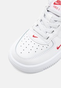 Valged Nike Air Force 1 ketsid sujuva nahast ülemise osaga, perforatsiooni saabastega, et tagada õhuringlus, ja punaste logo aktsentidega.
