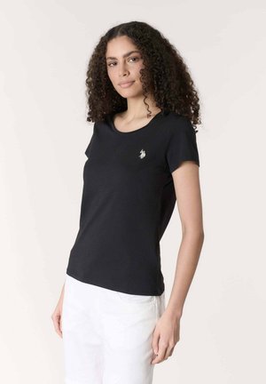 Donna con capelli ricci che indossa una maglietta nera a maniche corte con un piccolo logo bianco e pantaloni bianchi, in piedi davanti a uno sfondo semplice.