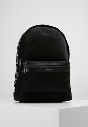 Musta Michael Kors -reppu, jossa on etutasku vetoketjulla, merkatut sivuhihnat ja minimalistinen muotoilu valkoisella jalustalla.