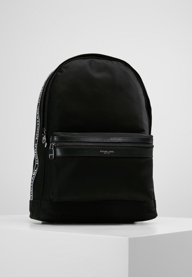 Sac à dos noir Michael Kors avec poche avant zippée, sangles latérales brandées et design minimaliste, posé sur un socle blanc.