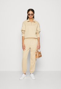 Beige quarter-zip sweatshirt with rolled sleeves, matching joggers, and white sneakers. A small brown handbag complements the outfit.