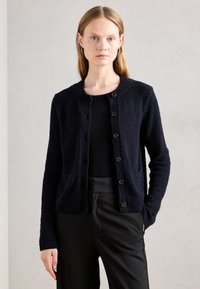 Frau mit glattem blonden Haar trägt eine schwarze, geknöpfte Strickjacke über einem schwarzen Top und schwarzen Hosen vor einem schlichten hellgrauen Hintergrund.