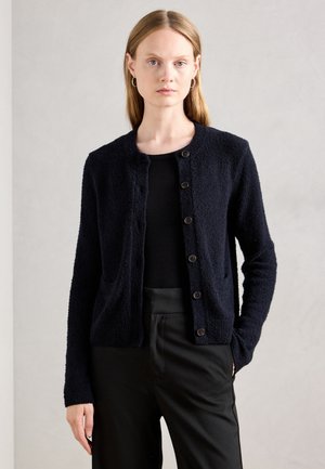 Vrouw met steil blond haar draagt een zwarte gebuttonde cardigan over een zwarte top en zwarte broek voor een effen lichtgrijze achtergrond.