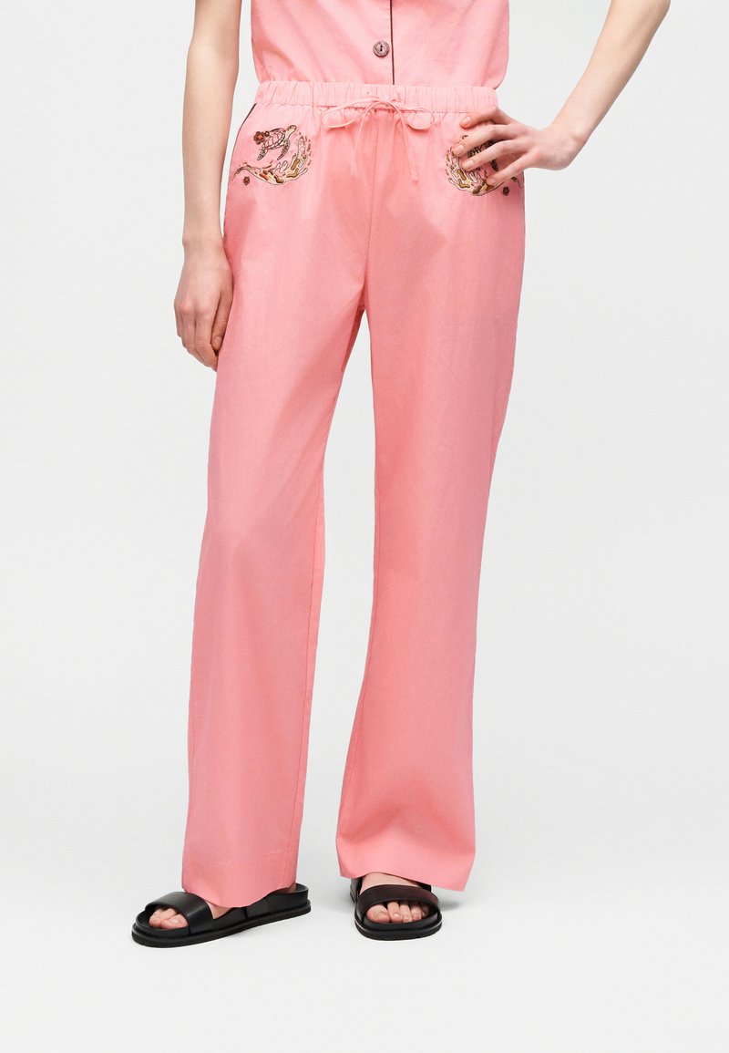 Luźne różowe spodnie z kwiatowym haftem na kieszeniach, z wiązanym w pasie sznurkiem, noszone z czarnymi sandałami na otwartym palcu, ręka modelki spoczywa na biodrze.