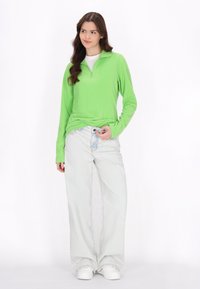 Pulóver de forro polar verde con cuello de media cremallera, combinado con pantalones vaqueros de pierna ancha en azul claro y zapatillas blancas. Texturas suaves y ajuste casual.