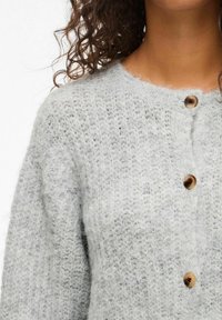 Ljust grå stickad cardigan med en mjuk, lurvig textur. Har rund halsringning och tre träknappar framtill.