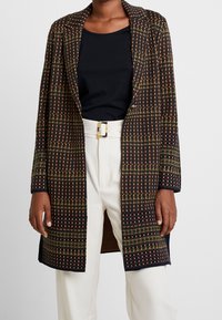 Manteau long à carreaux en tricot noir, orange et vert, avec un col à revers, une fermeture ouverte et des poches latérales. Porté sur un haut noir et un pantalon blanc.