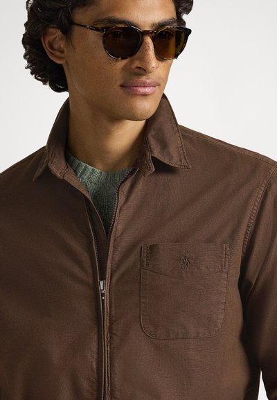 Καφέ μπουφάν με φερμουάρ �και γιακά, με μπροστινή τσέπη και διακριτική λεπτομέρεια λογότυπου. Φορεμένο πάνω από πράσινο πουλόβερ και συνδυασμένο με στρογγυλά γυαλιά ηλίου.