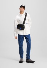 Crni crossbody torba s plavim akcentima, pravokutnog oblika, glatke teksture, postavljena na modelu koji nosi bijelu majicu i plave traperice.