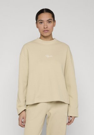 Kvinde med mørkt hår iført en beige ribbet langærmet top og matchende bukser, stående foran en ensfarvet lys baggrund.