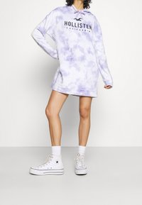 Mikina s kapucí v odstínu světle fialové s tie-dye, s potiskem loga. V kombinaci s bílými vysokými teniskami a ponožkami až po kotníky.