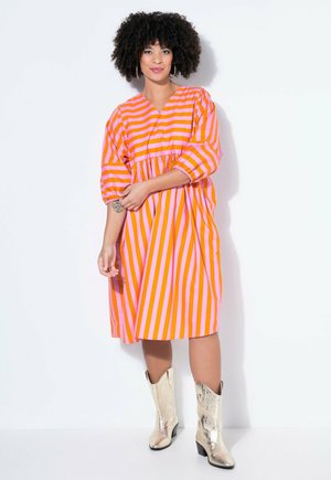 Mujer con cabello rizado vistiendo un vestido a rayas naranja y rosa y botas vaqueras metálicas plateadas, de pie contra un fondo blanco.