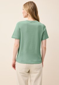 Camiseta de algodón verde con cuello redondo y mangas cortas, que presenta un corte holgado y una textura suave, combinada con pantalones beige claros.