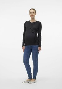 Maglietta premaman nera a maniche lunghe con dettaglio a nodo frontale, abbinata a jeans skinny blu e scarpe flat argentate. Look semplice e informale.