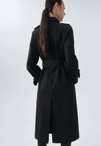 Trench coat negro con cinturón en la cintura, mangas largas y epaulets. Presenta una abertura en la parte posterior y una tela texturizada para mayor durabilidad.