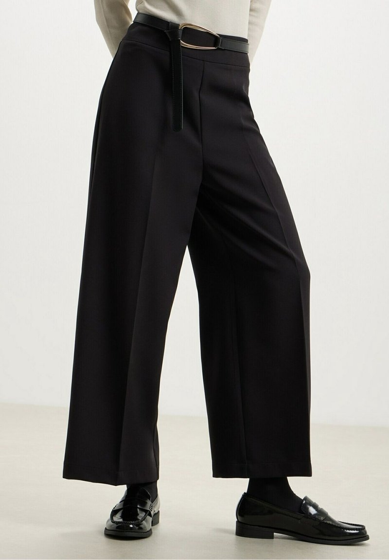 Pantalon noir à jambes larges associé à une ceinture noire, des mocassins noirs et un haut blanc, présenté de la taille aux pieds sur un fond uni.