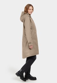 Beige knielange jas met capuchon, zijzakken en knoopdetails. Gedragen over donkere jeans en zwarte stevige laarzen. Glad stoffen textuur.