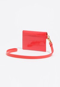 Czerwony portfel na karty z lakierowanej skóry z wzorem aligatora, wyposażony w pasek i złocisty klips. Kompaktowy i płaski design.