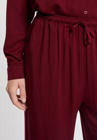 Close-up van een persoon die donkerrode elastische broek met een verstelbare trekkoord draagt, met een bijpassend langärmige top, hand ontspannen aan de zijkant.