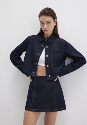 Dunkelblaue Jeansjacke mit silbernen Knöpfen und Brusttasche, paired mit einem passenden Jeansrock. Einfaches Design mit sichtbaren Nähdetails.