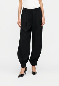 Γυναίκα που φοράει μαύρο παντελόνι ψηλής μέσης με στενή γραμμή και μανσέτες με κουμπιά στους αστραγάλους, καθώς και μαύρα μυτερά παπούτσια με ψηλό τακούνι, στέκεται μπροστά σε λευκό φόντο.