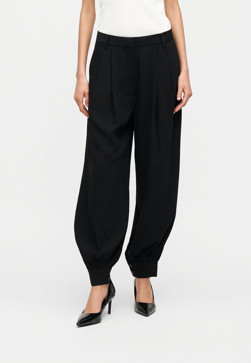 Γυναίκα που φοράει μαύρο παντελόνι ψηλής μέσης με στενή γραμμή και μανσέτες με κουμπιά στους αστραγάλους, καθώς και μαύρα μυτερά παπούτσια με ψηλό τακούνι, στέκεται μπροστά σε λευκό φόντο.