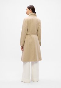 Abrigo de lana beige con un cuello alto, cintura ceñida y mangas largas, visto desde la parte de atrás. Pantalones de pierna ancha blancos visibles en la parte inferior.