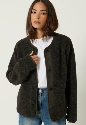 Femme portant une veste polaire sombre boutonnée sur une chemise blanche et un jean bleu, tenant la veste près du col.