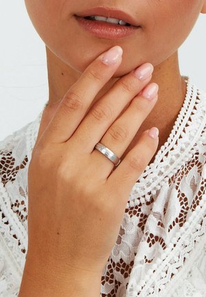 Sølvring med en glat, poleret overflade, båret på en hånd med lys rosa manicure, mod en baggrund af hvidt blonderstof.