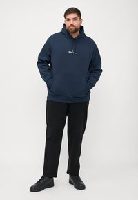 Felpa in cotone di colore blu navy con tasca frontale e cappuccio regolabile con lacci. Presenta un piccolo logo bianco sul petto. Abbinata a pantaloni e scarpe nere.