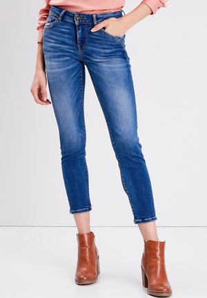 Vrouw draagt mid-rise skinny blauwe jeans en bruine enkellaarsjes met hak, met één hand in de zak en een roze shirt met lange mouwen.