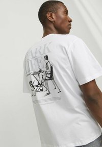 Hvid bomulds t-shirt med en sort grafisk illustration på bagsiden af en person ved et bord med en hund, ledsaget af tekst.