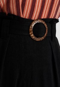 Black high-waisted trousers with a textured fabric. Features a brown circular belt buckle and pleats for added detail.