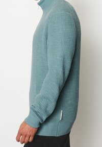 Pull-over en tricot côtelé vert d'eau clair avec col haut, manches longues et étiquette latérale, présentant un tissu texturé et une coupe ajustée.