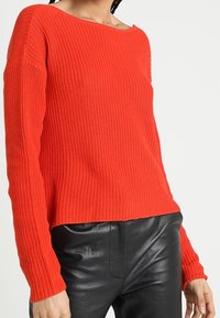 Czerwony sweter z dzianiny o szerokim dekolcie, długich rękawach i luźnym kroju. W zestawieniu z czarnymi skórzanymi spodniami o gładkiej fakturze.