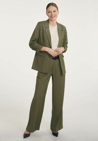Młoda kobieta w oliwkowym garniturze, białej koszuli, czarnym pasku i czarnych szpilkach, uśmiechnięta i stojąca na tle jednolitego, jasnego tła.