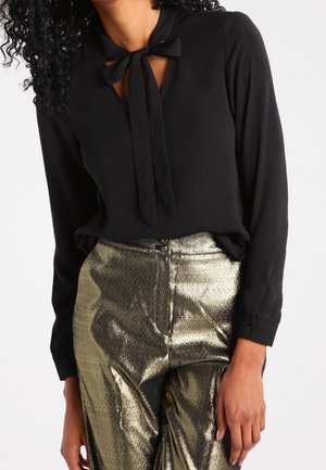 Kobieta ubrana w czarną bluzkę z długim rękawem z wiązaniem pod szyją i metaliczne złote spodnie z wysokim stanem.