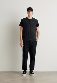 Crna quilted prsluk s patentnim zatvaračem, nošen preko crne majice i uparen s crnim hlačama i tenisicama. Jednostavan, monohromatski dizajn.