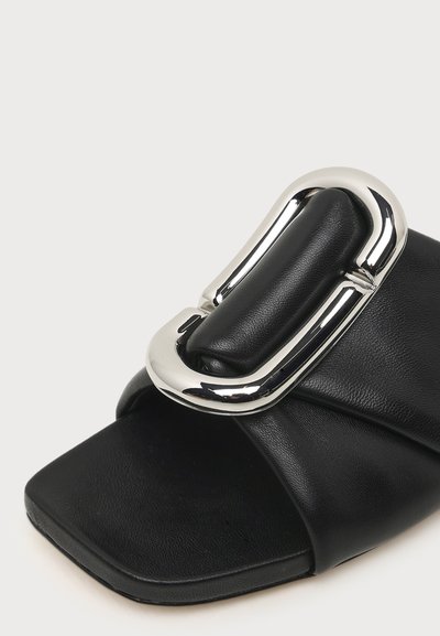 Sandale mule en cuir noir avec une grande boucle rectangulaire argentée et un design à bout carré sur un fond uni.