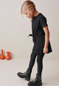 Sort tekstureret kjole med en bånd i taljen, parret med sorte leggings og sorte ankelstøvler. Baggrunden prydes af orange keramik.