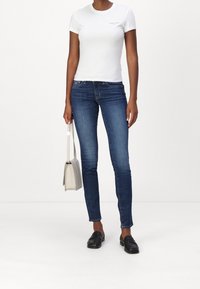 Camiseta blanca ajustada con logo, jeans ajustados azul oscuro, mocasines negros y un bolso gris claro. Diseño simple y casual.