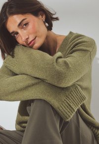 Žalias megztinis su V formos iškirpte ir grioveliais rankogaliais, derinamas su pilkomis kelnėmis. Minkšta tekstūra, laisvas siluetas. Minimalistinis dizainas.