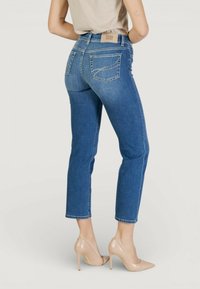 Femeie purtând blugi albaștri slim-fit cu detalii din cristale pe buzunarele din spate și pantofi nude cu toc înalt și vârf ascuțit, stând cu spatele întors.