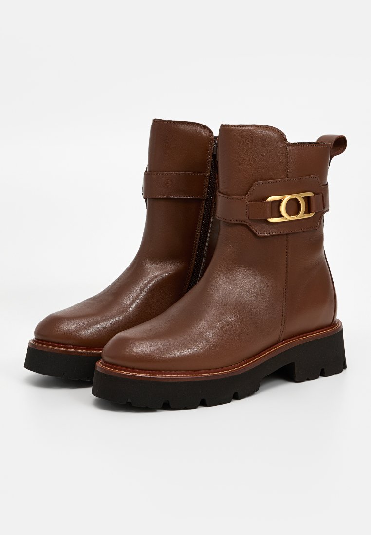 Braune Leder-Stiefeletten mit einem seitlichen Reißverschluss, grober schwarzer Sohle und einem goldenen, runden Schnallenakzent an einem Riemen. Glatte Textur, sauberes Design.