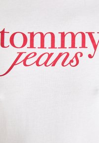 Baltos medvilnės marškinėliai su ryškiu raudonu "tommy jeans" užrašu stilizuotu šriftu krūtinės srityje, glotni tekstūra, neformalus dizainas.