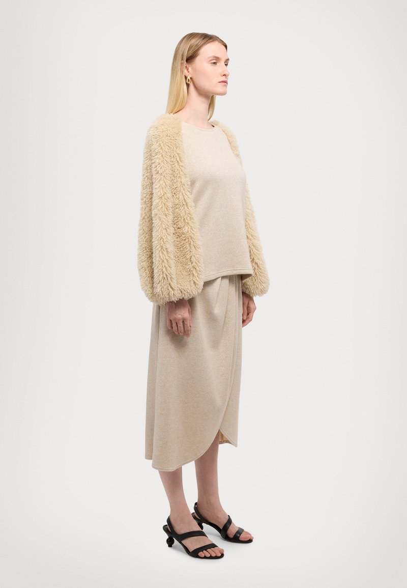 Beige Strickpullover mit flauschigen Ärmeln, kombiniert mit einem beigefarbenen drapierten Rock und schwarzen Riemchensandalen. Glatte Textur, gemütliches Design.