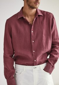 Bordowa lniana koszula zapinana na guziki z długimi rękawami, z szerokim kołnierzem i białymi guzikami, zestawiona z jasnymi spodniami.