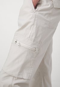 Pantalones cargo de tela beige claro, con un bolsillo con cremallera en el lateral, superficie texturizada y detalles de costura reforzada.